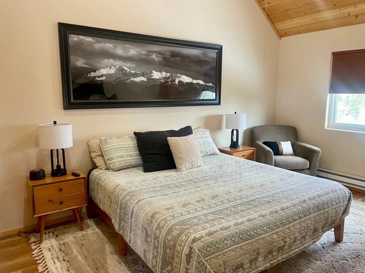 A spacious and inviting king bedroom featuring a large bed with soft, neutral linens, an armchair for reading, nightstands with glowing lamps, and a mountain photograph above the headboard. Natural light streams in from the windows, offering glimpses of the forest outside.
