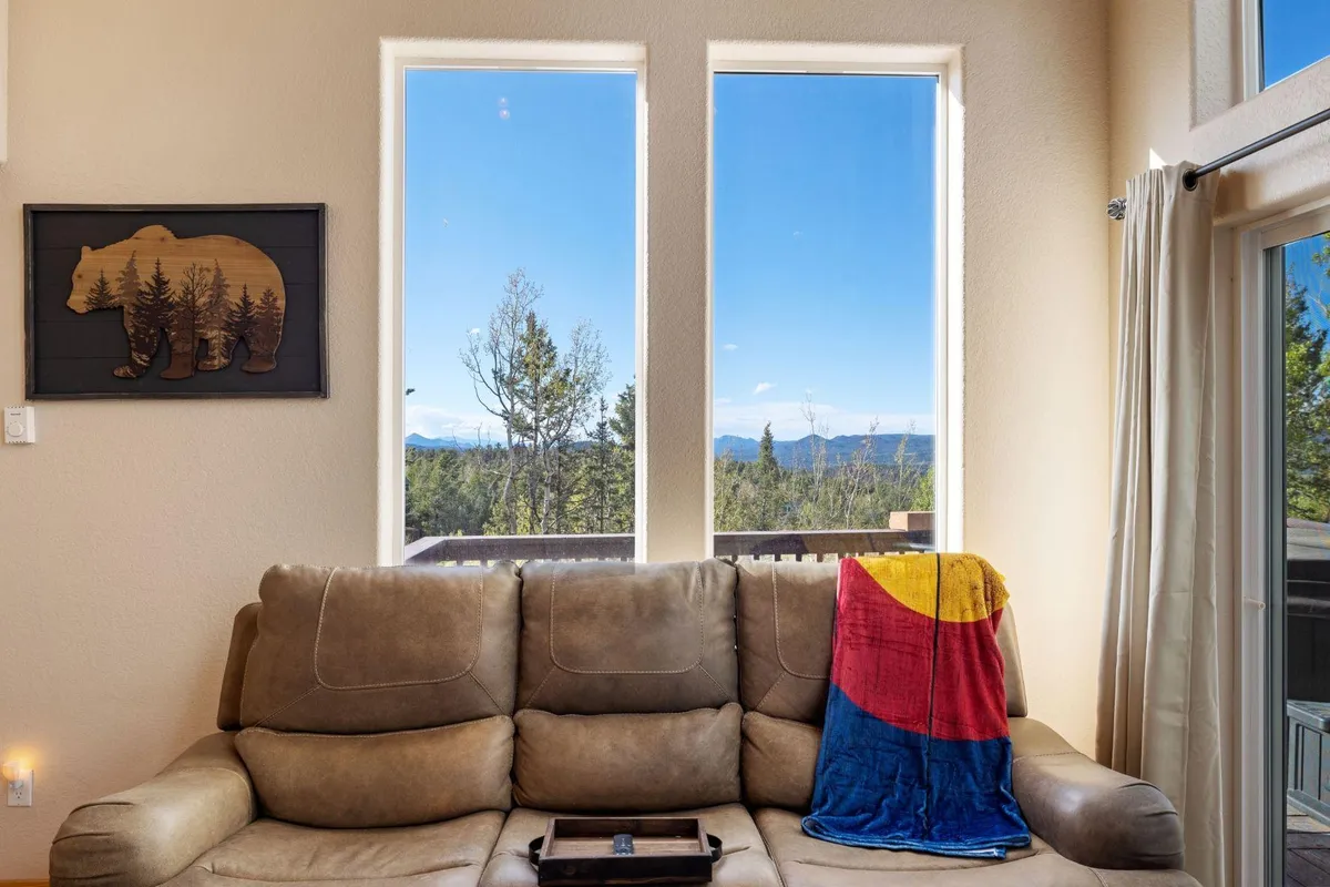 A comfortable living area with a beige sofa, colorful blanket, and decorative bear wall art. Large windows open up to mountain views, creating a warm and welcoming environment ideal for socializing, reading, or observing nature from indoors.