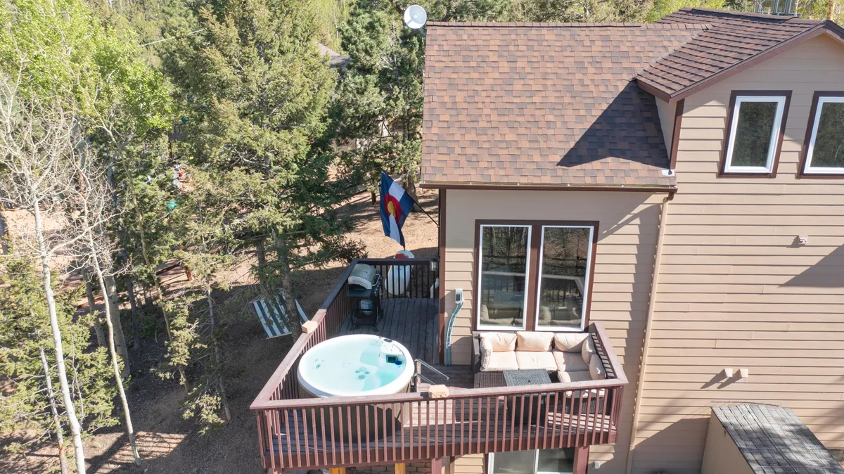 The image features a private hot tub situated on an outdoor deck, accompanied by comfortable seating and framed by a tree line. A Colorado flag adds a sense of place, while soft sunlight filters through the branches, creating a cozy, intimate atmosphere. The hot tub is the focal point, inviting viewers to imagine soaking while taking in the peaceful surroundings.