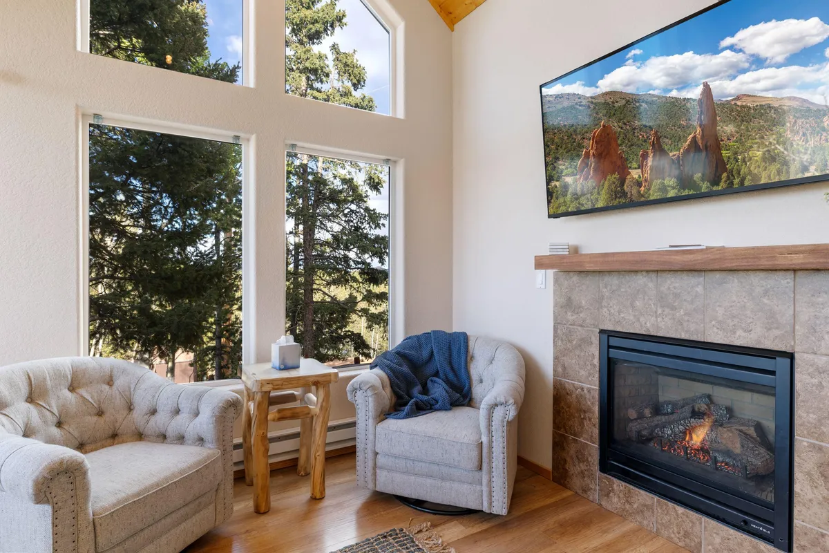 A spacious living area featuring large windows that frame breathtaking mountain views. Comfortable armchairs are arranged around a warm fireplace, with a television mounted on the wall. The ambiance is both inviting and cozy, making it perfect for relaxation, socializing, or enjoying the scenery.