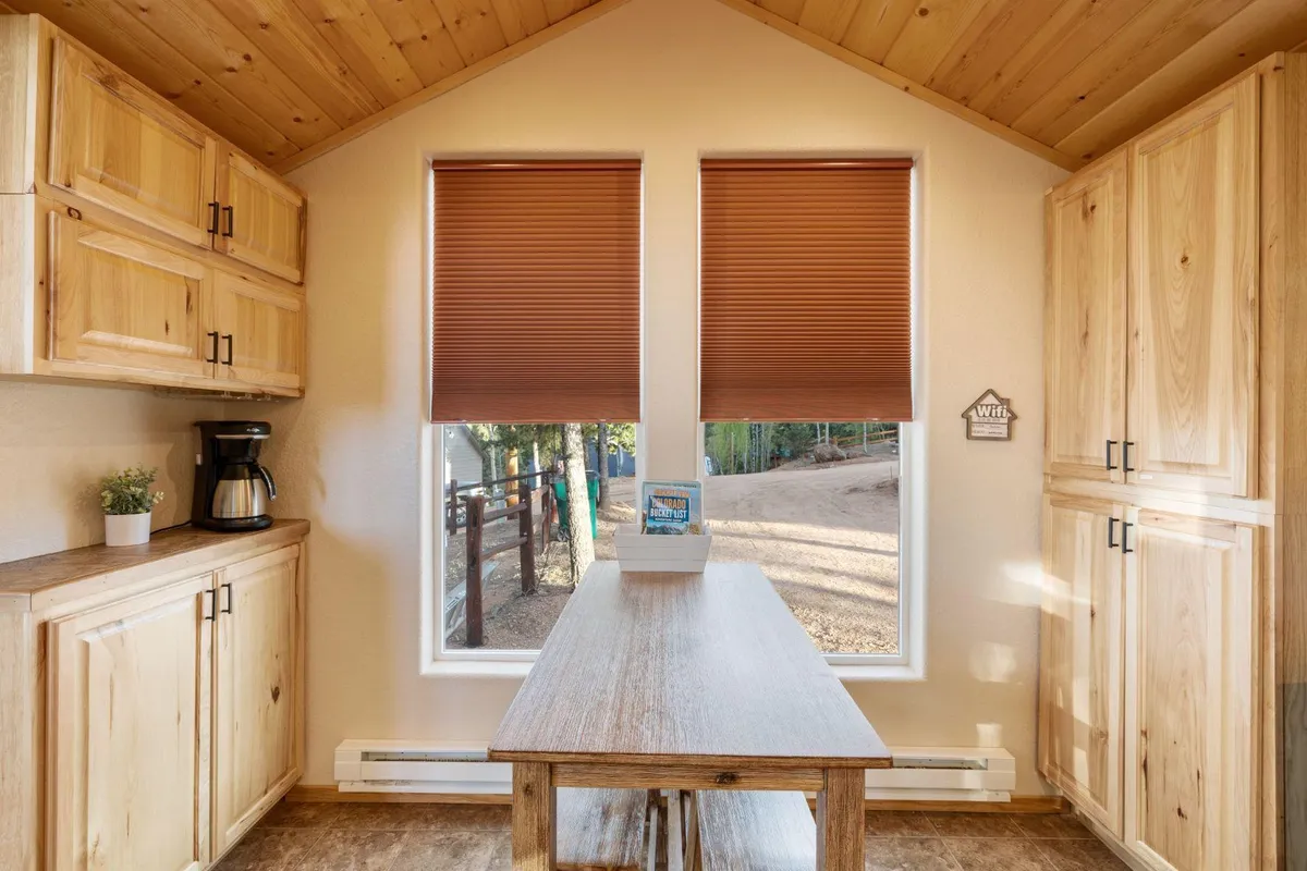 A cozy dining space with a wooden table positioned near large windows, natural wood cabinetry, and soft, ambient daylight streaming in. The space captures an inviting, communal atmosphere ideal for breakfasts and family dinners.