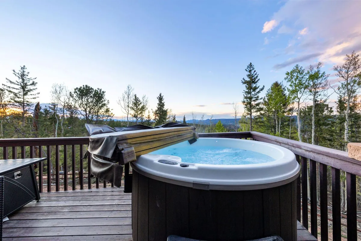 An inviting outdoor deck features a bubbling hot tub positioned to overlook a sweeping mountain vista. Comfortable seating is arranged next to the tub, and the deck is framed by wooden railings and surrounded by lush trees, creating an intimate connection to nature.