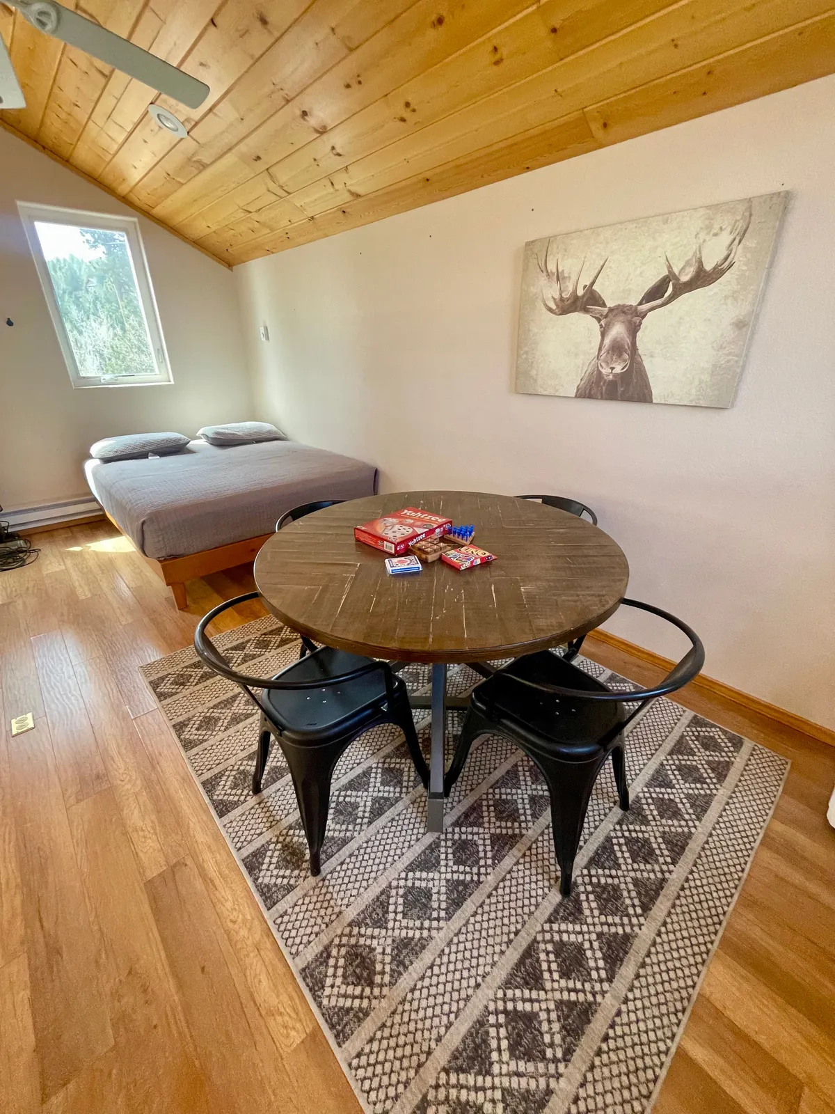 An open, airy interior view capturing a wooden ceiling, a round dining table, and a bed with neutral bedding. Moose artwork adds to the cozy, mountain-lodge feel. The perspective suggests the presence of a lofted area nearby.